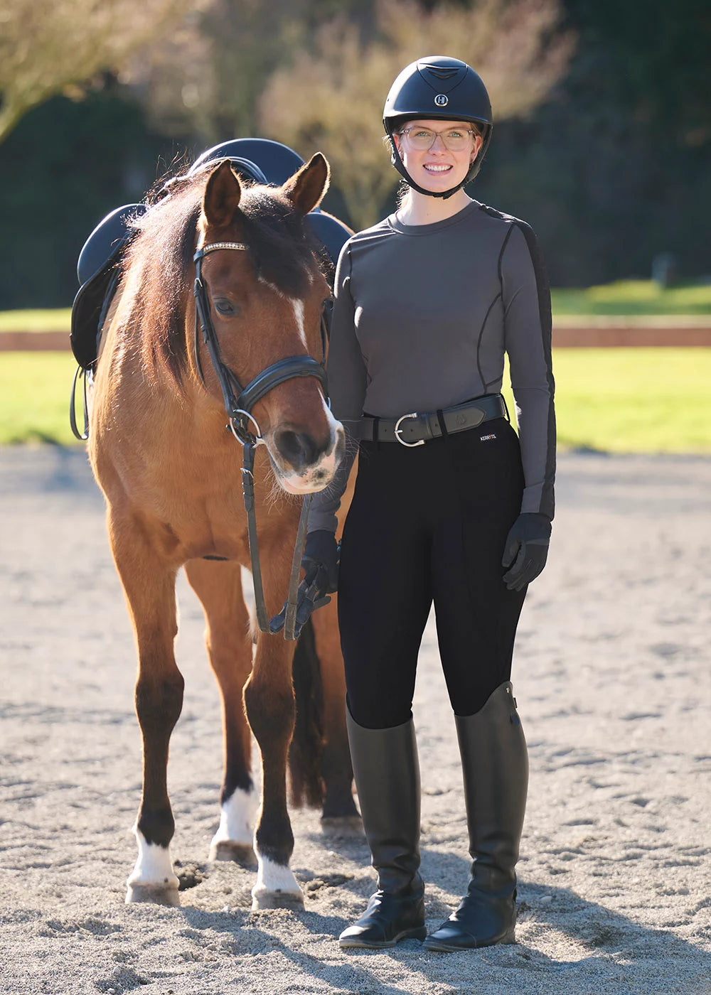 Front view of a female model wearing the Carbon Kerrits Bit of Base Layer Top while standing next to a horse in an outdoor arena