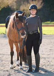 Front view of a female model wearing the Carbon Kerrits Bit of Base Layer Top while standing next to a horse in an outdoor arena
