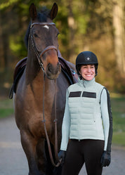 Front view of a female model wearing the Shoreline Kerrits Bit of Base Layer Top while standing in front of a dark colored horse