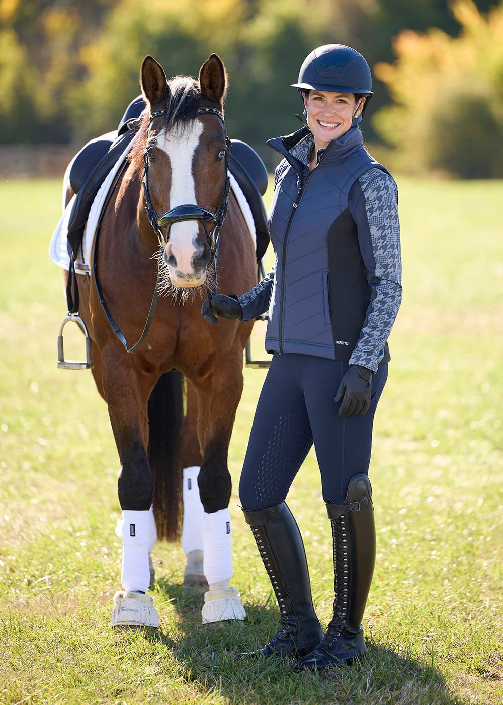 Angled front view of a female model wearing the Dusty Blue Kerrits Traverse Quarter Zip Fleece Tech Top under a coordinating vest while standing next to a bay horse outside
