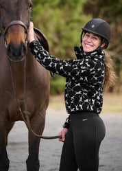 Side view of a female model wearing the Black Kerrits Berber Flying Horse Jacket while standing next to a bay horse outside