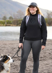 Front view of a female model wearing the Black Kerrits Cozy Crest Plush Fleece Sweatshirt while standing on a riverbed with a dog on a leash