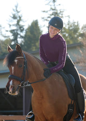 Front view of a female model wearing the Elderberry/Sugarplum Kerrits Equi Rhythm Crew Neck Sweater while riding a bay horse