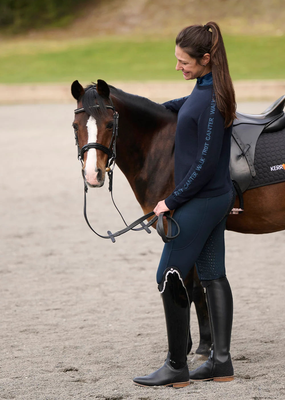 Side view of a female model wearing the Nightfall/Jasper Kerrits Equi Rhythm Crew Neck Sweater while standing next to a bay pony in an outdoor arena