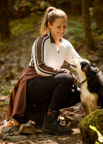 Woman wearing a light beige long sleeve riding shirt, squatting down to pet a dog outdoors 