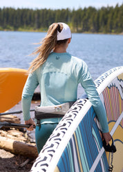 Back view of a female model wearing the light blue Kerrits Precision Long Sleeve Crew Neck Tech Top while carrying a paddle board