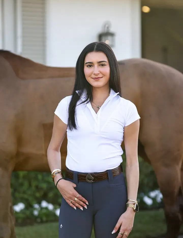 Front view of a female model wearing the white Criniere Margot Short Sleeve Show Shirt while standing in front of a bay horse