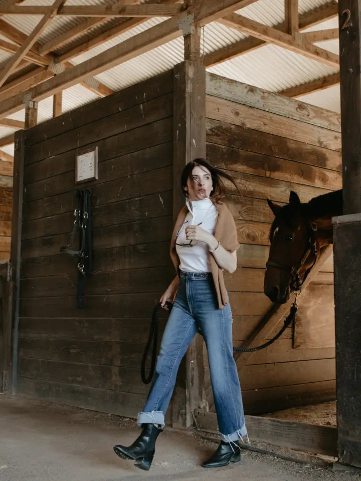 Angled front full body view of a female model wearing the white Criniere Margot Short Sleeve Show Shirt with jeans while waling a horse out of a stall