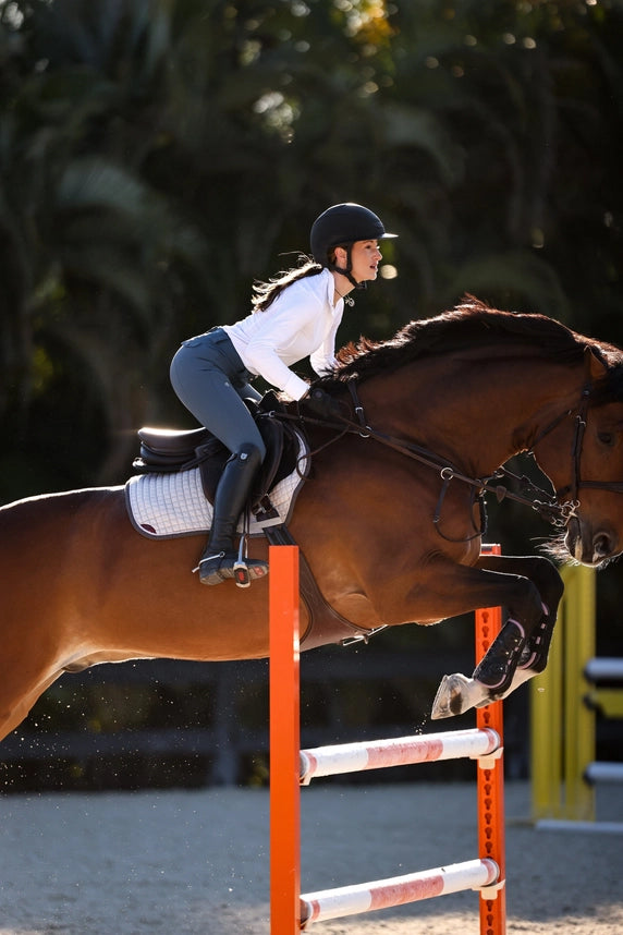 Side view of a female model wearing the white Criniere Helen Show Shirt while jumping a bay horse