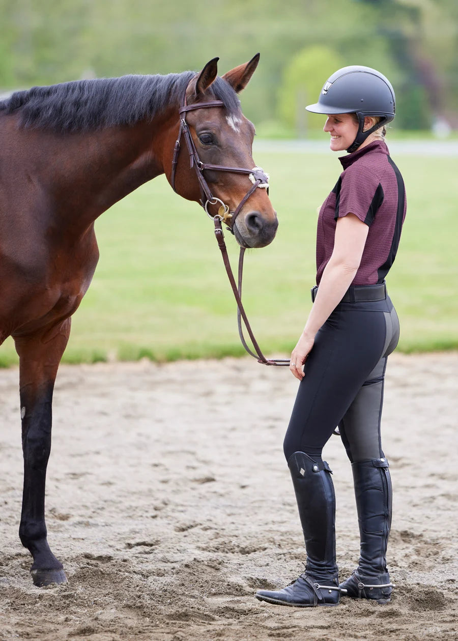 Side view of a female model wearing the black Kerrits Crossover II Full Seat Breech while standing in front of a horse in an outdoor arena