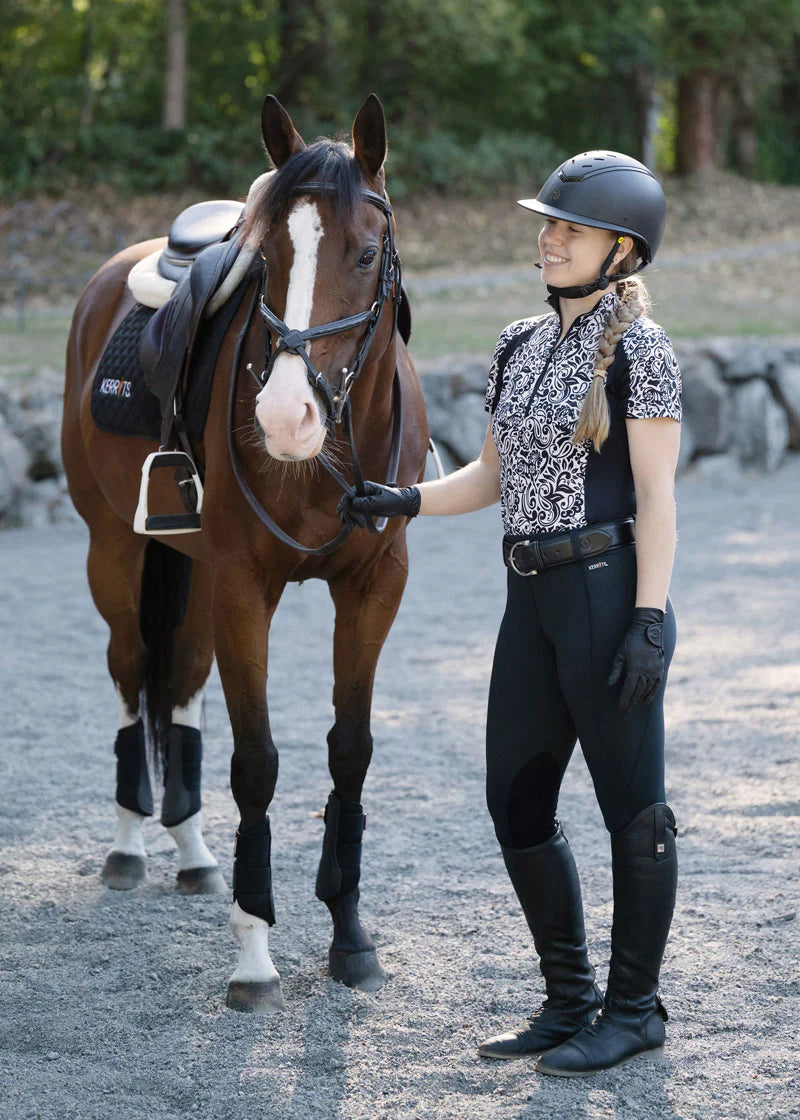 Full body front view of a female model wearing the black Kerrits Performance Knee Patch Pocket Tight while standing next to a horse