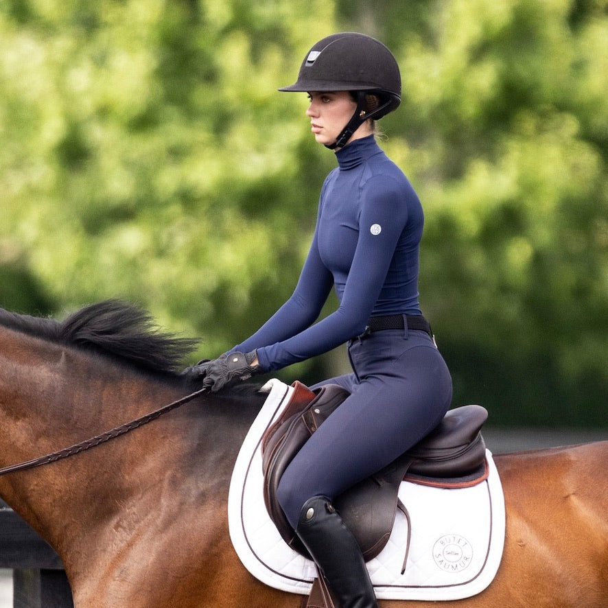 Side view of a female model wearing the Ink TKEQ Chloe High Collar Technical Top while riding a bay horse outside