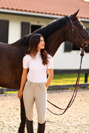 View of a female model wearing the tan Criniere Juliette High Compression Half Seat Show Breeches and a white shirt while standing in front of a dark colored horse