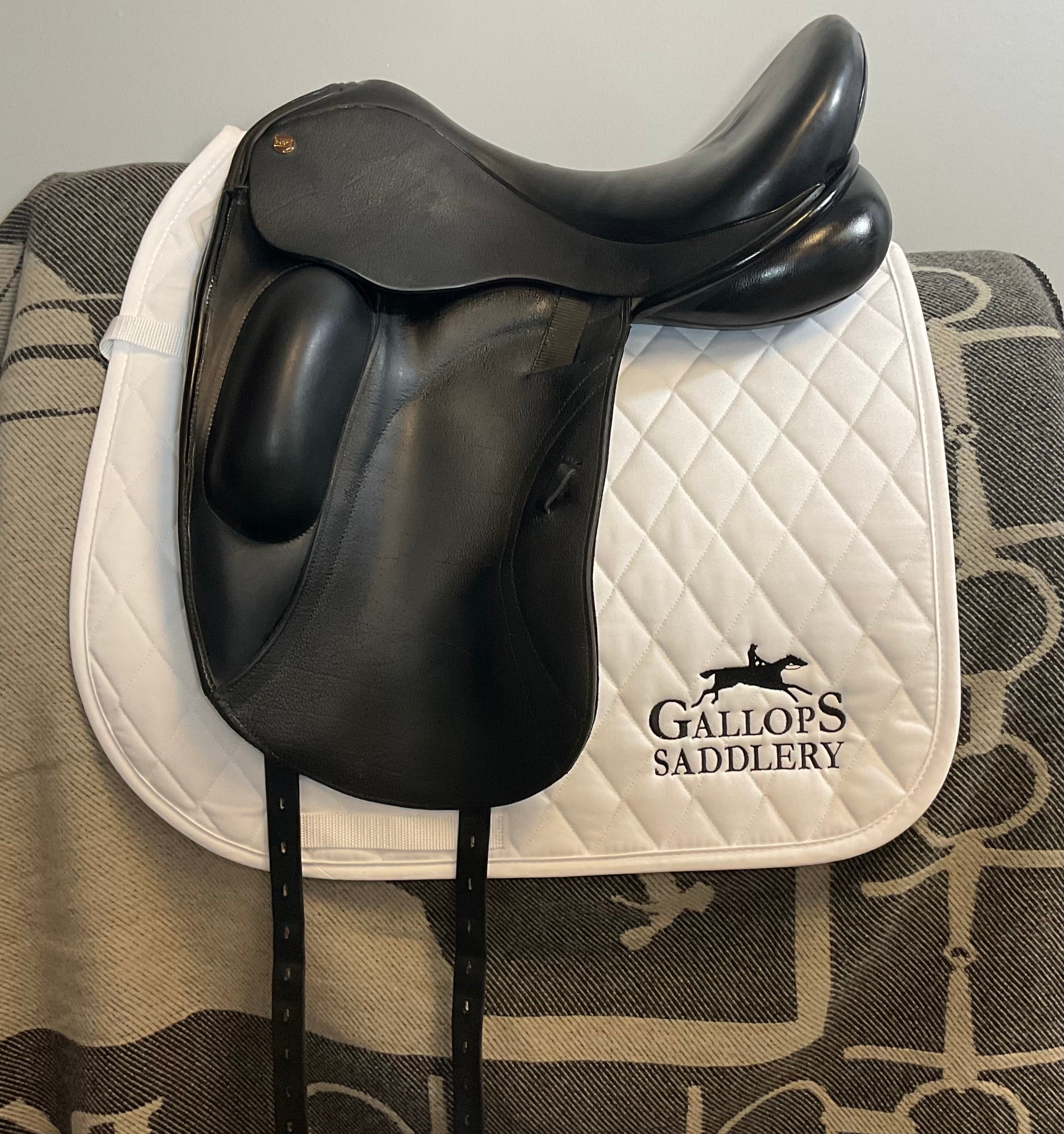 A black leather equestrian saddle resting on a white quilted saddle pad with the 'Gallops Saddlery' logo printed on it, set against a patterned gray and beige blanket.