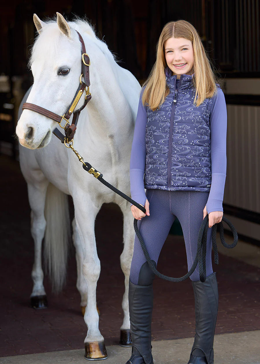 Young girl standing next to a white pony out of a barn while wearing the purple Kerrits Kids Fleece Lite II Knee Patch Tight and coordinating outfit