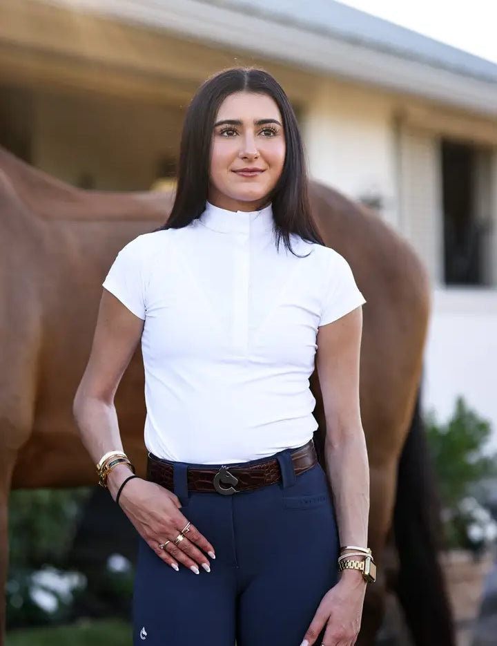Front view of a female model wearing the white Criniere Margot Short Sleeve Show Shirt while standing in front of a bay horse