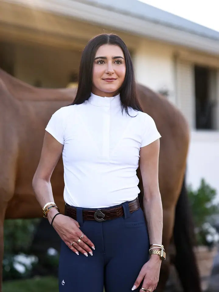 Front view of a female model wearing the white Criniere Margot Short Sleeve Show Shirt while standing in front of a bay horse
