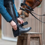 Model putting on a black Kavalkade "SafeHeel" Faux Fur Spur Protector with foot raised on wooden stool