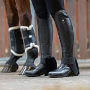 Side view of a model wearing the black Kavalkade "SafeHeel" Faux Fur Spur Protector over black tall boots while standing in a barn aisle