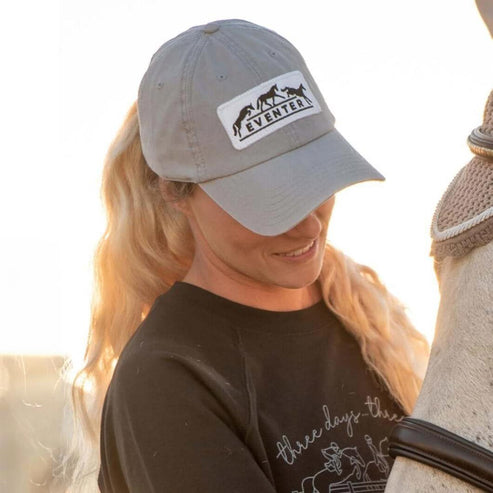 Angled front view of a model wearing the Dapplebay Eventer Barn Hat while standing next to a gray horse