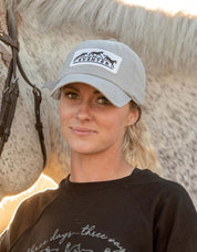 Front view of a female model wearing the Dapplebay Eventer Barn Hat while standing in front of a gray horse