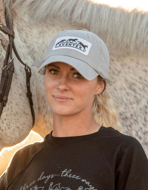Front view of a female model wearing the Dapplebay Eventer Barn Hat while standing in front of a gray horse