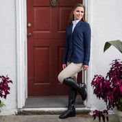Full body angled front view of a female rider wearing the tan Aubrion Suffolk Knee Patch Breeches with a blue show coat and black tall boots in front of a red door