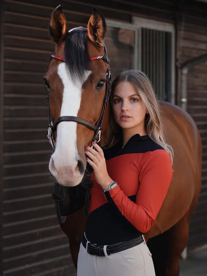 Front view of a bay horse and a female model who is wearing the black/red Criniere Sophie Fierce Focus Long Sleeve Shirt with a barn in the background