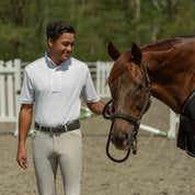 Front view of a man standing in an outdoor arena next to a horse while wearing the tan Equinavia Karl Men's Knee Patch Breeches and white shirt