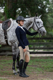 Angled front view of a model wearing the beige Free Ride PRO Knee Patch Breeches while standing in front of a gray horse