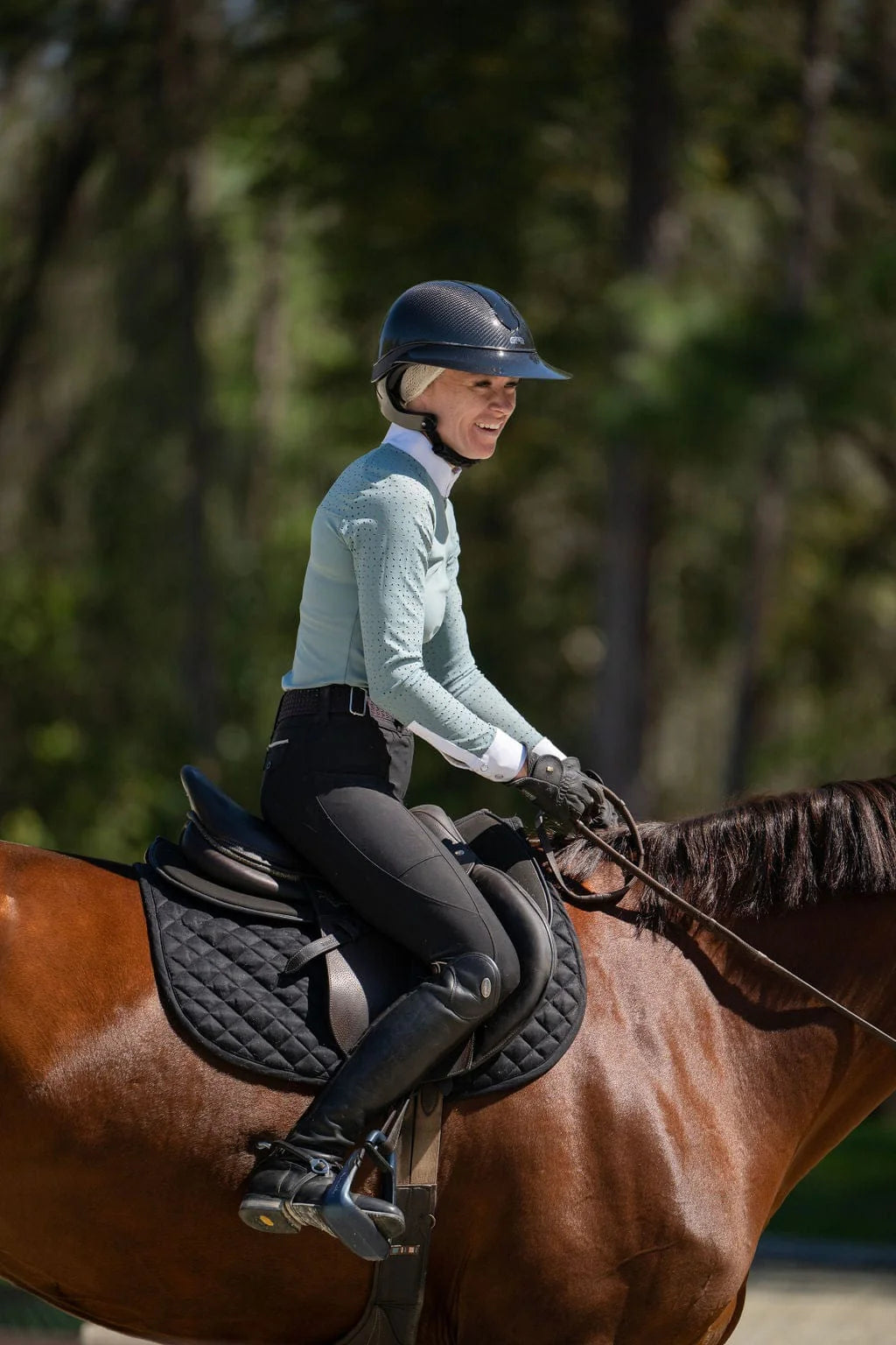 Side view of a female model wearing the black Free Ride Lux Knee Patch Breeches and green top while riding a bay horse