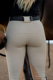 Back view of a model wearing the beige Free Ride PRO Knee Patch Breeches while standing in front of a dark colord horse in an outdoor arena