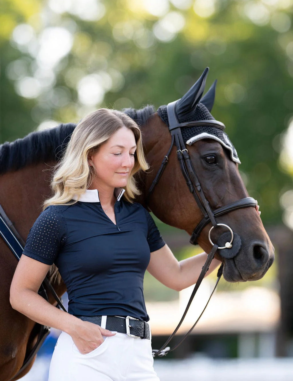 Angled front view of a female model wearing the navy Free Ride Cassidy Short Sleeve Show Shirt while standing next to a bay horse
