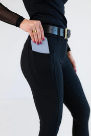 Side view of a model wearing the black Free Ride Sparkle PRO Full Seat Breeches and putting a phone in the pocket on a plain background