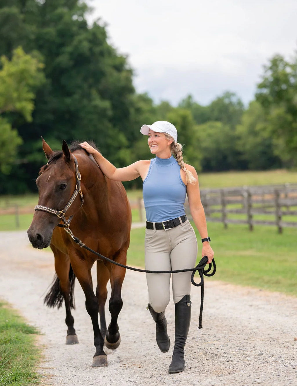 Front view of a woman wearing the beige Free Ride Sparkle PRO Full Seat Breeches while walking a bay horse on a gravel path