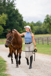 Front view of a woman wearing the beige Free Ride Sparkle PRO Full Seat Breeches while walking a bay horse on a gravel path