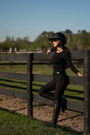 Angled front view of a model wearing the black Free Ride PRO Knee Patch Breeches and black shirt while leaning up against a black wooden fence