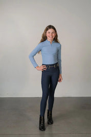 Full body front view of a young model wearing the navy Free Ride Youth Lux Full Seat Breeches and blue top on a plain background