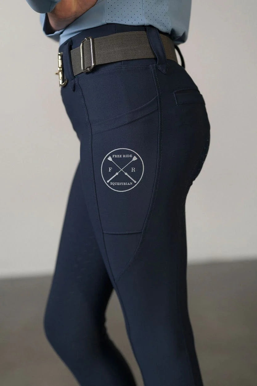 Close-up side view of a young model wearing the navy Free Ride Youth Lux Full Seat Breeches on a plain background