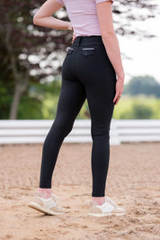 Angled back view of a female model wearing the black Free Ride Lux Knee Patch Breeches while standing in an outdoor arena