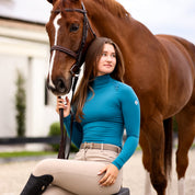 Front view of a female model wearing the Gulfstream TKEQ Chloe High Collar Technical Top while sitting in front of a chestnut horse