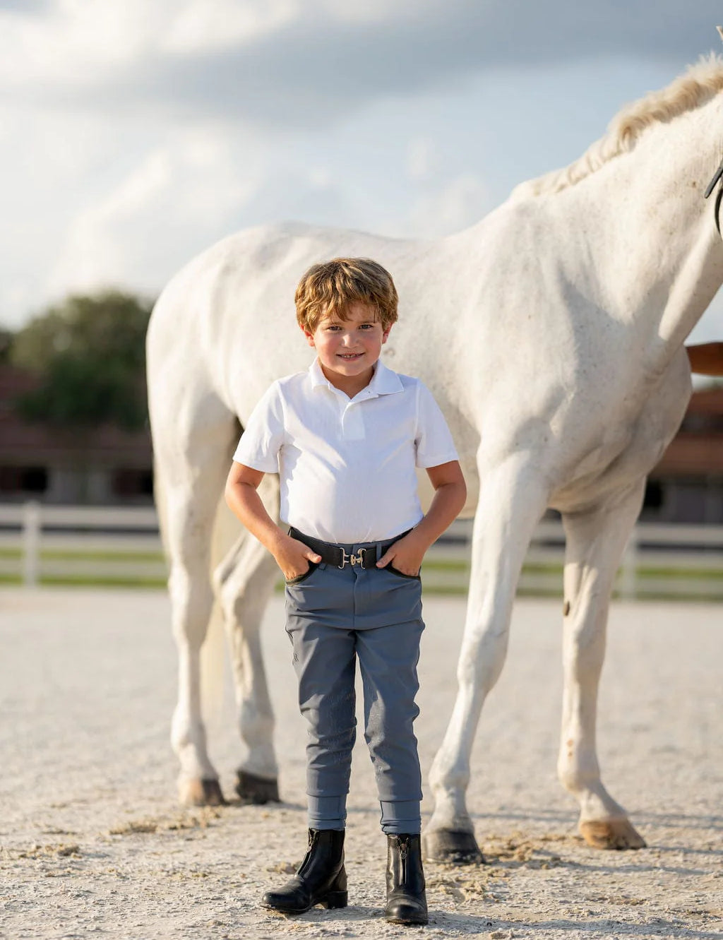Full body front view of a young male model wearing the blue Free Ride Boys Pro Knee Patch Breeches while standing in front of a gray horse