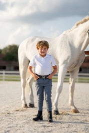 Full body front view of a young male model wearing the blue Free Ride Boys Pro Knee Patch Breeches while standing in front of a gray horse