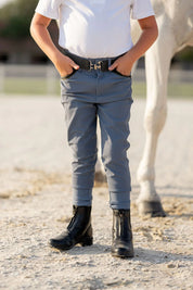 Front view of a young male model wearing the blue Free Ride Boys Pro Knee Patch Breeches while standing in front of a gray horse