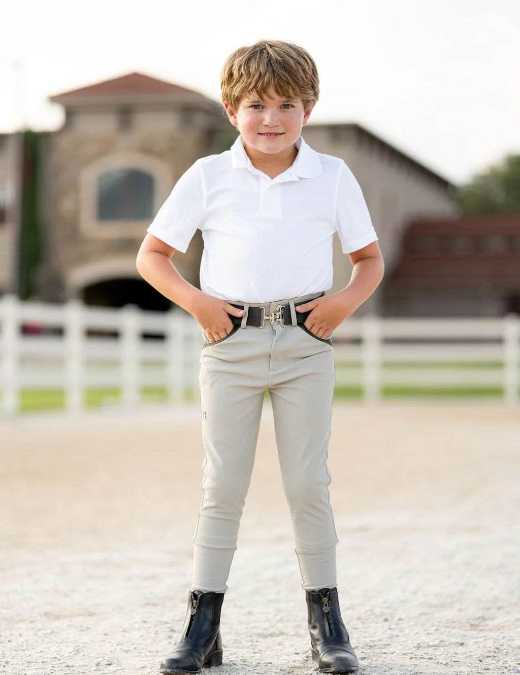 Full body front view of a young male model wearing the beige Free Ride Boys Pro Knee Patch Breeches and white shirt while standing in an outdoor arena