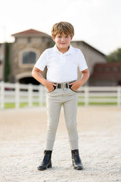 Full body front view of a young male model wearing the beige Free Ride Boys Pro Knee Patch Breeches and white shirt while standing in an outdoor arena
