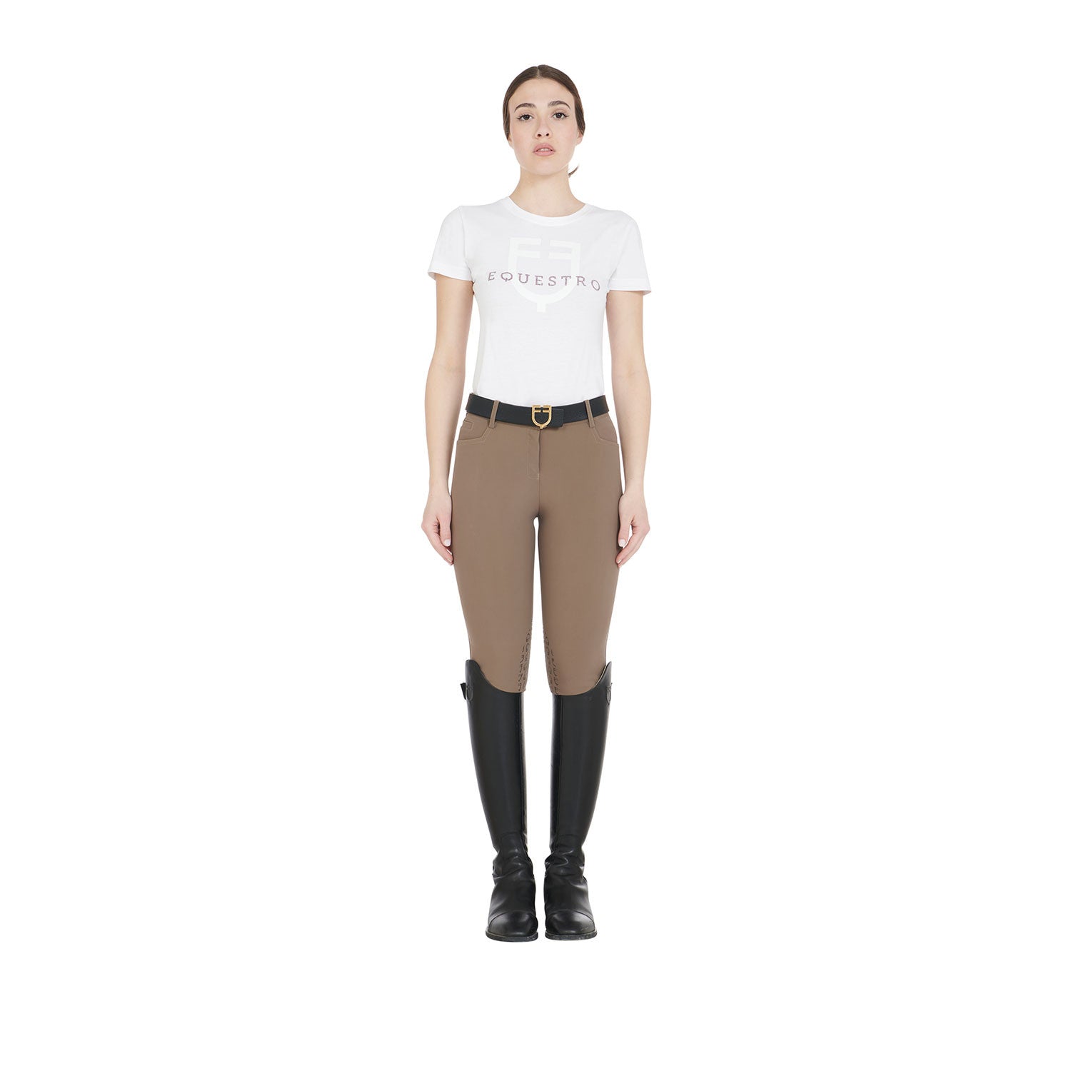 Full body view of a model wearing the Equestro Women's Slim Fit Grip Breeches With Logo in tan, white tshirt, and tall black boots on white background