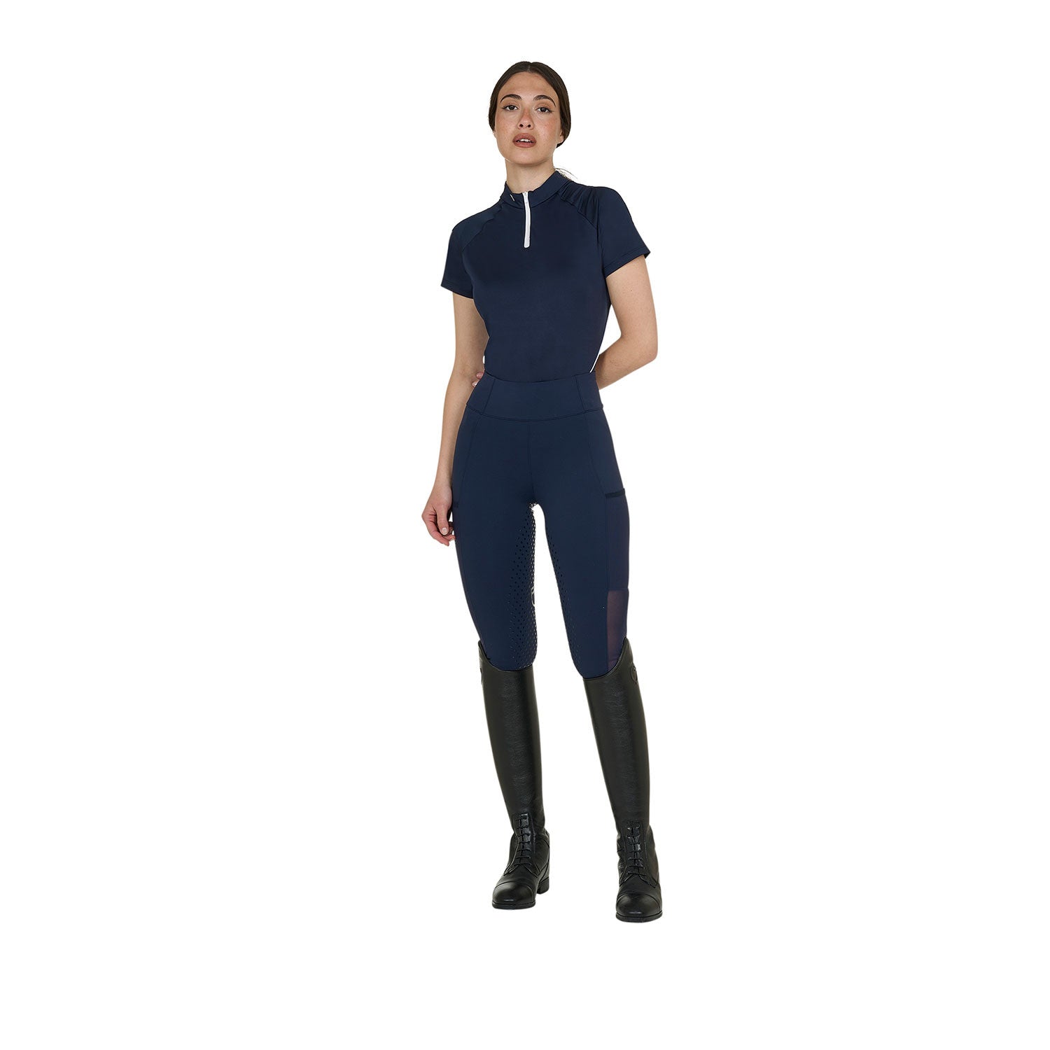 Bull bodied front view of a model wearing the Equestro Women's Slim Fit Technical Training Polo Shirt in navy with navy tights and black tall boots