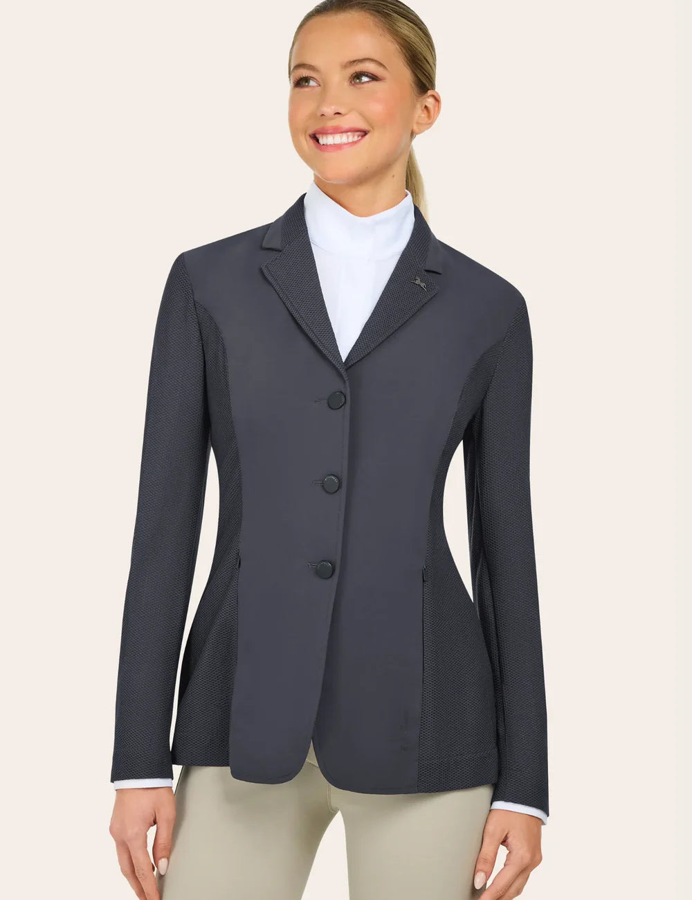 A woman wearing a dark gray tailored blazer over a white shirt and beige pants stands facing slightly to the side and smiles.