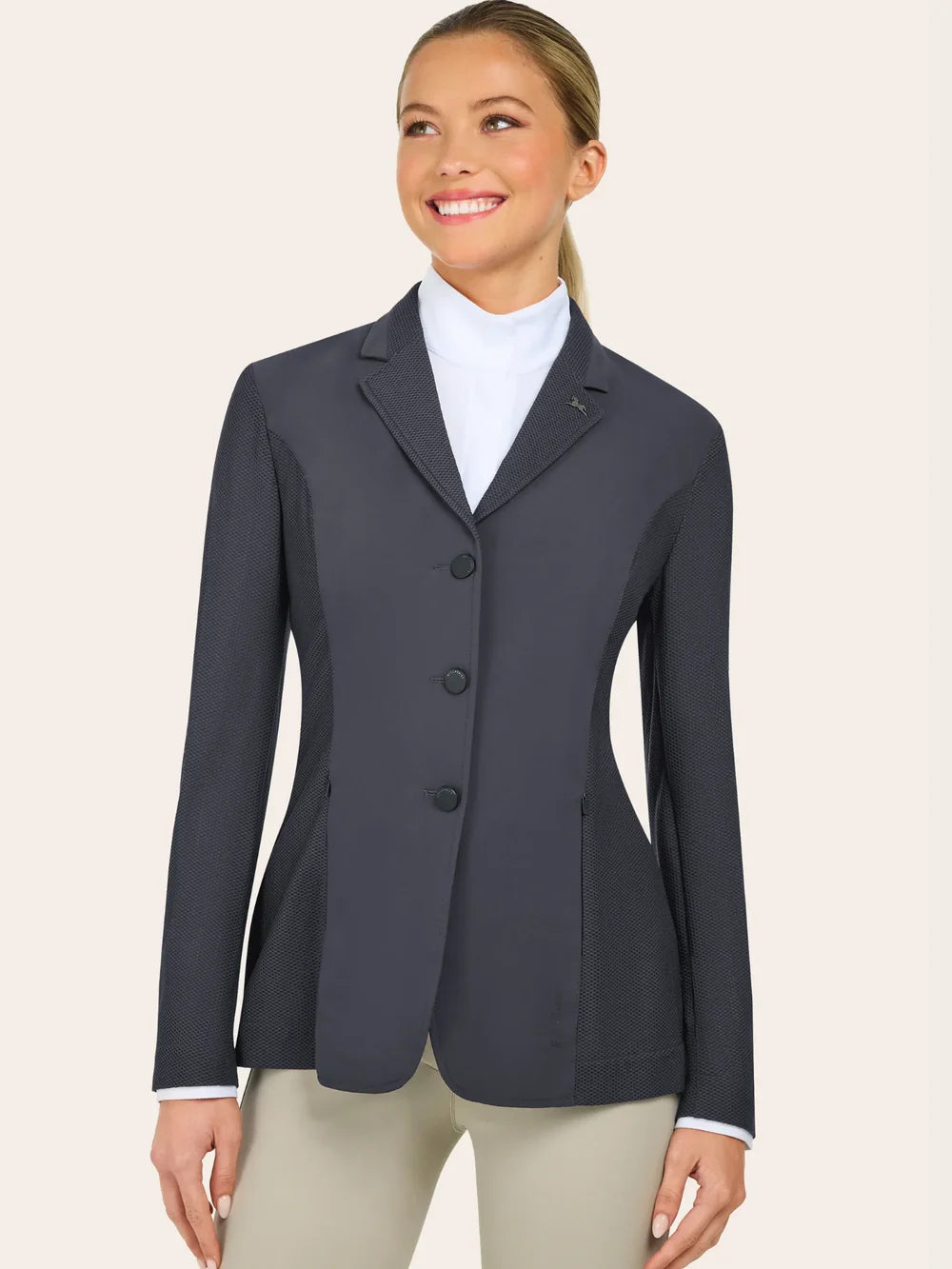 A woman wearing a dark gray tailored blazer over a white shirt and beige pants stands facing slightly to the side and smiles.
